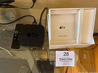 Apple TV 4K device with remote control and original box displayed on a table with logo and instructions card inside the box.