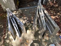 Two folded metal wheelchair ramps standing upright on a concrete surface outdoors with some leaves around.
