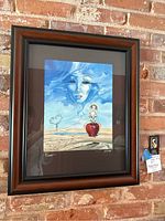 Framed artwork with surreal imagery featuring a female face in the sky, a girl on a red fruit, desert background.