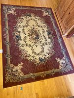 Full view of the rectangular area rug on a hardwood floor showing central floral design framed by ornate burgundy border with mixed color accents.