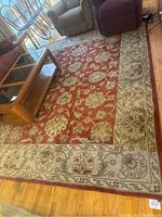 Full view of the large rectangular floor rug with red center and beige floral border, placed in living room with wooden coffee table and upholstered furniture around it.