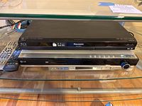 Two media players stacked on glass shelf: Panasonic black Blu-ray player on top, LG silver DVD player below, with LG remote next to them.