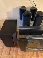 Six LG surround sound speakers displayed, including subwoofer, center speaker, and four satellite speakers on a glass table.