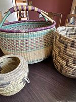 Three handwoven Native American baskets on a table, showing the large lidded basket with colorful handle, large open geometric pattern basket, and smaller rounded basket.
