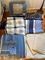 Photo showing a navy blue Goose Down comforter in sealed packaging next to several quilts and pillowcases laid out on the floor.