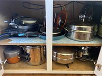 Photo showing various pots, pans, electric fryer, griddle, and other cookware stacked on shelves including pots with lids and pans with handles.