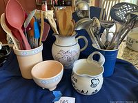 Full view of pottery pitchers, crocks, bowl and kitchen utensils including wooden and stainless utensils displayed on a blue cloth background.