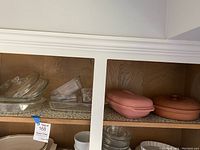 Wide view of shelving showing clear glass Pyrex bakeware on left, and pink Pic stoneware and Tupperware casserole dishes on right.