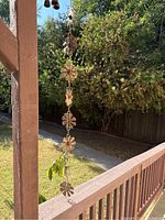 Vertical photo of wooden flower-shaped rain catcher and wind chime hanging from a wooden post, outdoors with surrounding greenery.