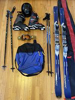 Photo showing black Giro helmet on top left, pair of Salomon ski boots with red accents, Oakley goggles, blue and black boot bag, two pairs of ski poles, and Atomic skis arranged on wooden floor.