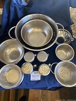 Set of large and small stainless steel colanders, five stainless bowls, and two sets of stainless steel salt and pepper shakers arranged on blue cloth.