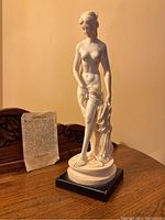 Front view of the white Venus La Baigneuse sculpture on black base with description paper in background