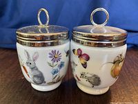 Front view of two vintage Royal Worcester porcelain egg coddlers with silver lids and loop handles, one featuring rabbit and floral decoration, and the other with mouse, hedgehog, butterfly, and fruit design.