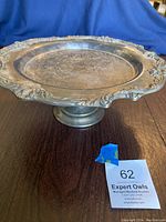 Front angle photo of vintage Reed & Barton silverplate cake plate on pedestal showing the raised ornate border and pedestal base on wooden table.