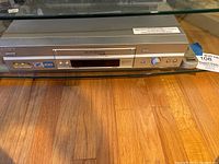 Front view of Sony VCR unit on wood floor, showing tape slot, buttons, knob, and RCA ports.