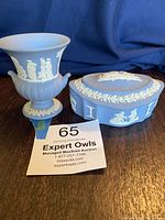 Photo showing two blue Wedgwood Jasperware pieces: an urn-shaped vase with classical white relief figures and a matching trinket box. Both displayed on a wooden surface with blue fabric backdrop.