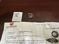 Photo of the ring laid on a dark wood surface alongside a certificate of appraisal and a lot number tag.