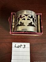 Close-up image of silver napkin ring inside pink-lined box, showing decorative cut-outs and size scale reference with handwritten note.