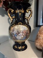 Front view showing the cobalt blue vase with gold handles and ornate gold detailing surrounding a classical painted scene of a family in 18th-century clothing.