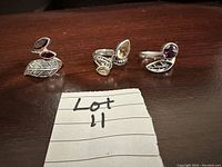 Three silver rings with garnet, topaz, and amethyst stones in leaf-themed settings shown on wood surface; Lot 11 label visible