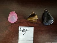 Three gemstone pendants displayed on a dark wood surface. Left to right: pink translucent, Tiger's Eye brown and gold, and dark gray with white veining. A handwritten note labeled 'Lot 7' is included.