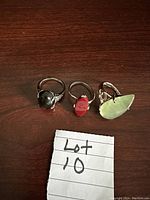 Three silver rings with solitaire gemstones in black, red, and pale green colors on a dark wood surface, with a note saying Lot 10.