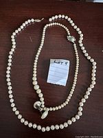Two faux pearl necklaces and a pair of faux pearl screw-back earrings laid out on a wooden surface with a handwritten lot number tag
