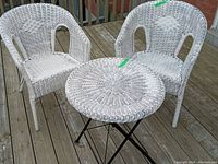 Two painted white wicker chairs and one round wicker table with black metal base on wooden deck outdoor.