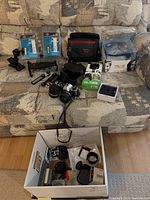 Photo showing Olympus OM10 camera with attached Soligor 28mm lens, Nikon 50mm lens, Soligor 75-200mm zoom lens, camera flash, photo case and various accessories on couch.