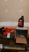 Image showing orange Remington electric chainsaw and metal animal live trap side by side on a shelf, with a bottle of chainsaw oil on top of the trap box.