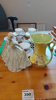 Full view of the yellow floral pitcher, porcelain figurine, and set of china teacups and saucers on table