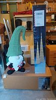 Photo showing boxed Noma outdoor slim Michigan pine tree and lighted gnome ornament with striped tall green and white hat, red and green outfit.