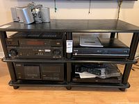 Four electronic components on a black shelf rack: top left four small speakers, top right Pioneer CD player, bottom left Toshiba DVD player, bottom right Sony BluRay DVD player with remote and manuals.