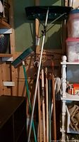 Full view showing the snow roof rake, broom, rakes, pitchfork, mop handles, and plastic garbage can in garage setting