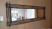 Front angled view of the forged metal framed wall mirror hanging inside a home, showing the crisscross metal design and reflections of a window and furniture.