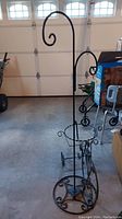 Full view of the metal plant stand inside a garage, showing the tall vertical pole with curved hook and some wheels and other objects in the background.