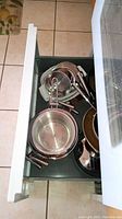 Drawer containing multiple stainless steel pots and pans including lids with metal handles, small and large sizes.