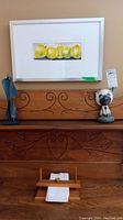 Framed watercolor print of yellow and green fruits by S. Happer hung above carved wood furniture with a blue steel sculpture on left, a carved wood animal figurine on right, and wooden napkin holder below