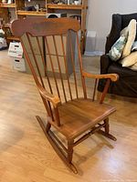 Wooden rocking chair shown at an angle, displaying the left side with a curved top backrest, vertical spindles, armrests, seat, legs, and rockers.