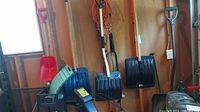 Three black plastic snow shovels with wooden handles hanging on a wall in a garage, with cords and other tools in the background.