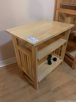 Angle view of the light wood side table showing top surface, two shelves, slatted sides, and casters placed on bottom shelf.