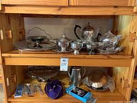 Wide view of wooden shelf displaying various silver plated tea and coffee pots, serving trays, covered serving dishes, and other silverware items including boxed accessories and purple pouch of spoons.