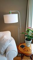 Metal floor lamp with brushed metal frame and white fabric drum shade next to a white couch and potted plant.