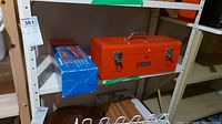 Orange metal Sears Craftsman tool box on shelf next to blue Eurofiles tile cutter box