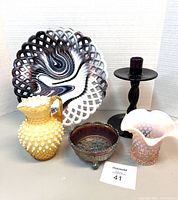 Full view of the lot including slag glass plate, yellow hobnail syrup jug, carnival glass butter dish, pink hobnail vase, and black candle holder.