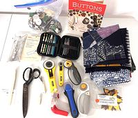 Overview of full craft lot showing rotary cutters, scissors, crochet hooks in case, fabric yards, buttons in package and bag, candle, and small utility knife.