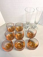 Photo of eight amber tinted hand blown glass with rounded bottoms and two tall clear pilsner glasses against white background.