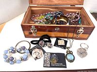 Costume jewelry stored in an open wooden jewelry box, including bracelets, earrings, pendants, and necklaces arranged outside and inside the box
