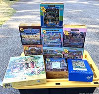 Nine jigsaw puzzles stacked on a yellow tray, showing puzzle box art and piece counts: seven puzzles are 500 pieces, two are 1000 pieces. Brands include Dowdle and Puzzazz.
