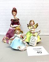 Three Josef Original figurines displayed together showing a woman in purple dress, child angel in blue dress holding a floral number 3, and an angel in yellow dress holding a doll.